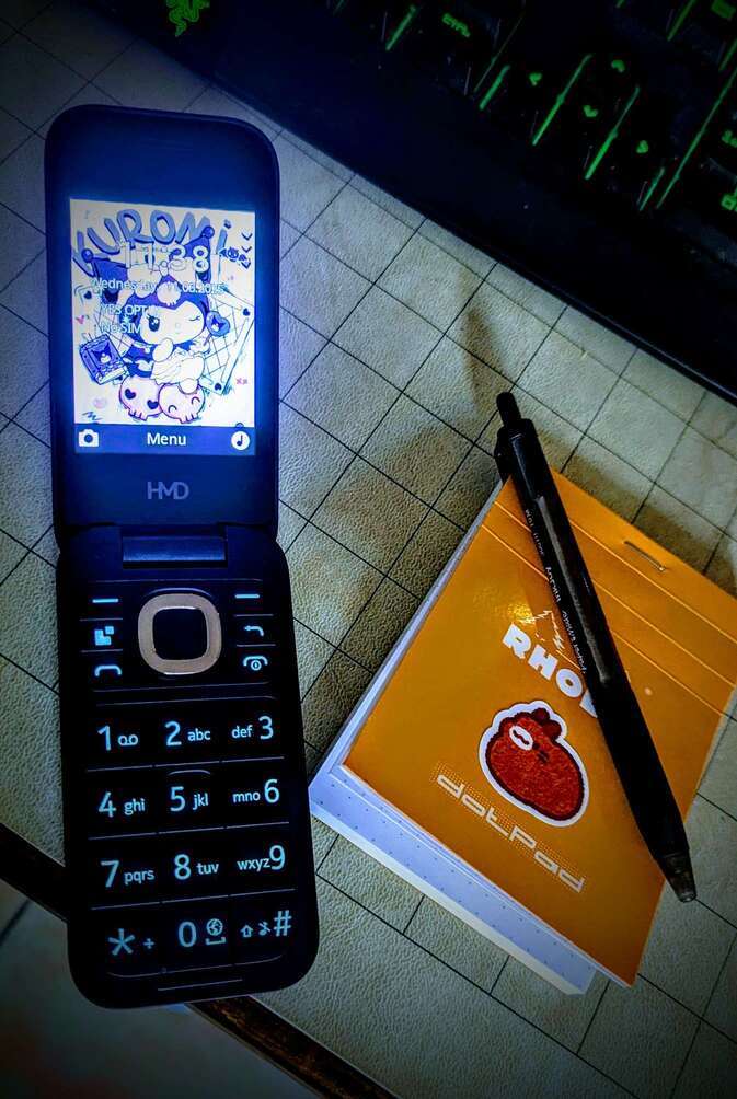 opened flip phone with numberpad and a kuromi wallpaper on the screen sitting next to an orange rhodia pocket dot pad with a pen. there's a cure red bunny sticker on the pad :)